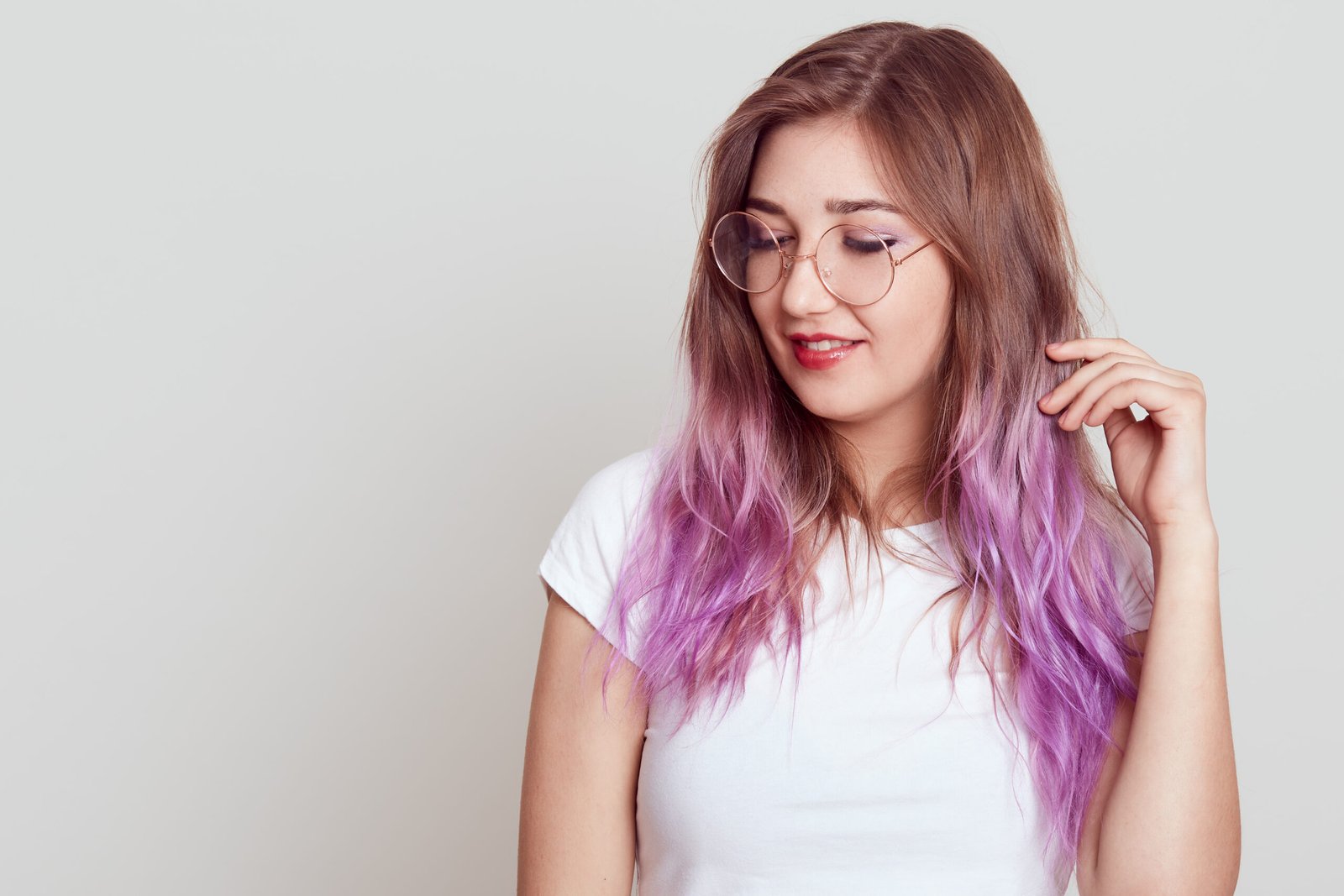 Dreamy attractive woman with lilac hair looking away and touching her hair, wearing white t shirt and glasses, copy space for advertisement, posing isolated over gray background.